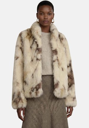 Femme portant une veste en fausse fourrure beige et marron tachetée, un pull beige, une jupe marron texturée et des boucles d'oreilles créoles dorées sur fond blanc.
