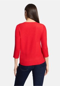 Czerwony, teksturowany sweter z dzianiny z rękawami do połowy przedramienia, okrągłym dekoltem i lekko zaokrąglonym dołem, zestawiony z ciemnoniebieskimi dżinsami.