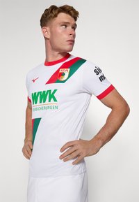 Hvit sportsdrakt med røde og grønne skråstriper, en rund rød krage, og logoen til FC Augsburg. Inkluderer sponsortekst i grønt.