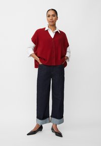 Gilet rosso oversize con bottoni su una camicia bianca, abbinato a pantaloni in denim scuro a gamba ampia con orli risvoltati e ballerine nere a punta.