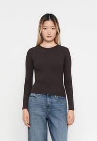 Ung kvinne med langt rett hår iført en tettsittende mørkebrun langermet skjorte og løse blå jeans, stående mot en ensfarget hvit bakgrunn.