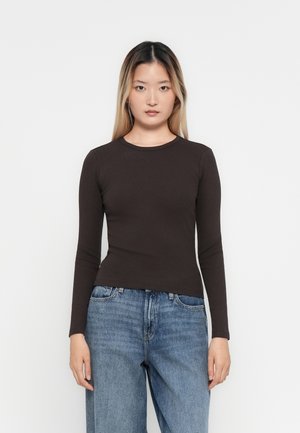 Ung kvinne med langt rett hår iført en tettsittende mørkebrun langermet skjorte og løse blå jeans, stående mot en ensfarget hvit bakgrunn.