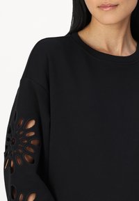 Black sweatshirt with cut-out floral pattern detail on sleeves. Soft fabric, crew neck design, and relaxed fit.