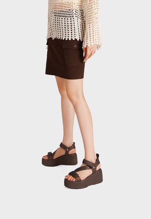 Donna con sandali platform marroni con cinturini alla caviglia, gonna marrone scuro con tasca laterale e top beige all'uncinetto.