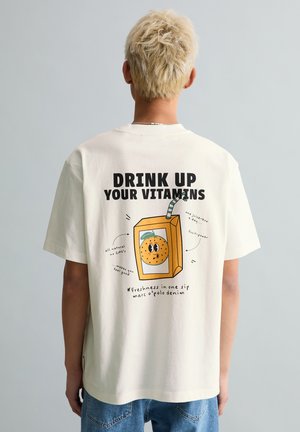 Personne aux cheveux courts décolorés portant un t-shirt blanc avec une illustration d'une brique de jus d'orange cartoon et le texte « Bois tes vitamines » au dos.
