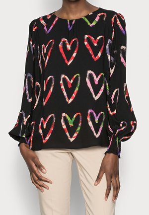 Black long-sleeve blouse with colorful, hand-drawn heart patterns worn with beige pants on a person standing.