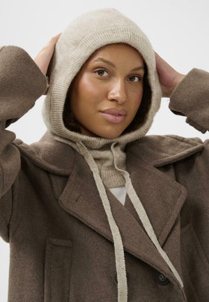 Brauner Wollmantel mit breiten Revers, darunter ein beige- und cremefarbener Kapuzenpullover. Die Textur ist weich, mit sichtbaren Nähdetails.