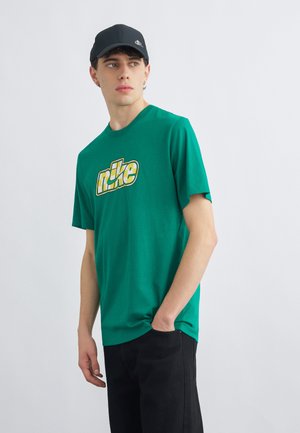 Junger Mann trägt ein grünes Grafik-T-Shirt mit gelbem und weißem Muster, schwarze Kappe und schwarze Hose, schaut zur Seite, eine Hand in der Tasche.