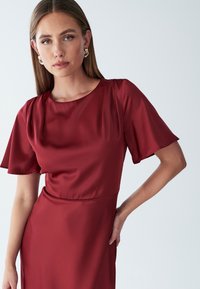 Bordo satenska obleka s kratkimi rokavi, okroglo izrezom in gladko teksturo. Opičena po svojem prileganju in subtilnih gubah na ramenskem delu.
