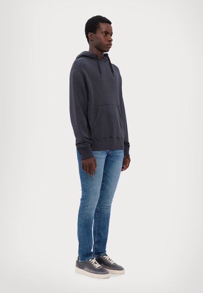 Tmavě šedá mikina s kapucí a přední kapsou, spárovaná se světle modrými slim-fit džíny a šedými teniskami s bílou podrážkou. Jednoduchý design, měkká textura.