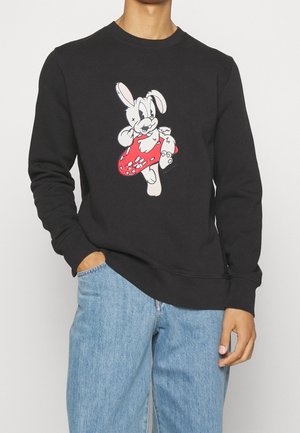 Personne portant un sweat-shirt noir avec un lapin blanc de dessin animé tenant un champignon rouge, associé à un jean bleu clair.