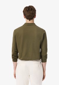 Olivenfarbener Langarm-Polohemd mit Kragen und hochgeschlagenen Ärmeln, kombiniert mit hellen Hosen, die ein einfaches Design und eine glatte Textur aufweisen.