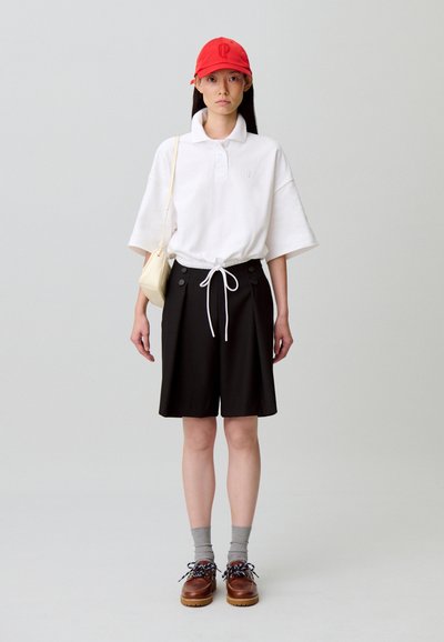 Polo blanc à manches courtes avec logo, short noir avec détails à boutons, casquette rouge, chaussettes grises, et chaussures marron à lacets. Design minimaliste.