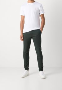 Mann mit den Händen in den Taschen, der ein weißes Kurzarm-T-Shirt, dunkle enge Hosen und weiße Sneakers trägt, vor einem einfarbigen Hintergrund.