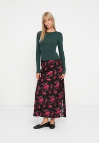 Grøn, ribbet langærmet top parret med en sort, blomsterprint maxi-nederdel med pink og røde blomster. Model iført sorte flade sko og en simpel frisure.
