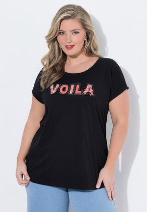 Donna bionda che indossa una maglietta nera con la scritta "VOILA" in lettere floreali rosa, abbinata a jeans azzurri chiari, in piedi davanti a uno sfondo bianco.