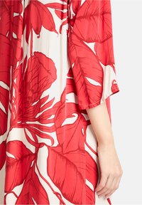 Rode bloemenpatroonkleding in een lichtgewicht materiaal. Bevat grote rode bloemen en bladeren op een lichte achtergrond. Loszittende mouwdetails zichtbaar.