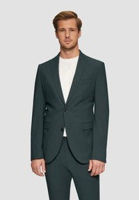 Dunkelgrüner maßgeschneiderter Blazer mit schlanker Passform, einreihiger Knopfverschluss, eingelegten Revers und zwei Vordertaschen. Kombiniert mit einem weißen Hemd.