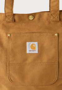 Καφέ τσάντα χειρός από καμβά με τσέπη, με πλακέτα λογότυπου Carhartt και δύο λουριά ώμου, με κούμπωμα τύπου κουμπί στο κέντρο πάνω μέρος.
