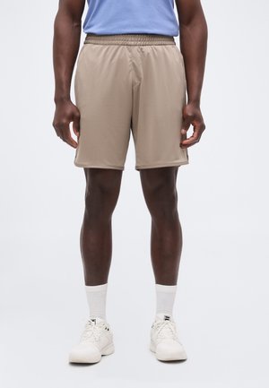 Mężczyzna ubrany w beżowe szorty z elastycznym pasem, jasnoniebieską koszulę, białe skarpety i białe trampki stoi na jednolitym tle.