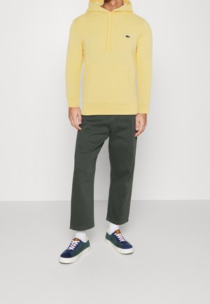 Homme portant un sweat à capuche jaune avec une poche frontale, un pantalon court vert foncé, des chaussettes blanches et des baskets bleues à semelles blanches.