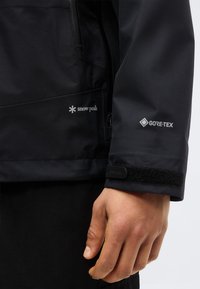 Gros plan de la main d'une personne et de la manche d'une veste noire arborant les logos Snow Peak et GORE-TEX avec un réglage du poignet en velcro.