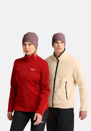 Frau in roter Fleecejacke mit Reißverschluss und schwarzer Hose und Mann in cremefarbener Fleecejacke und schwarzer Hose, beide tragen lila Mützen.
