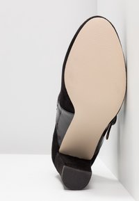 Zapato de tacón negro con textura de ante suave, acento de charol brillante y suela de goma beige que presenta un patrón de agarre texturizado.