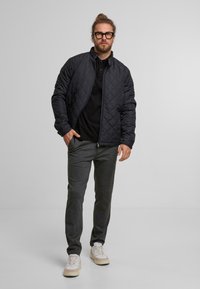 Czarny pikowany kurtka z stójką, noszona na czarnej koszulce polo. Szare zwężane spodnie i jasne sneakersy dopełniają outfit.