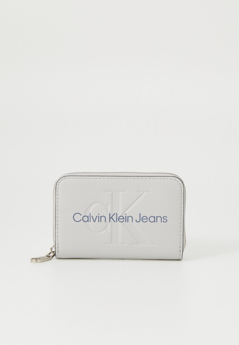 Pilkas odinis piniginė su iškilmu "CK" logotipu ir mėlynu "Calvin Klein Jeans" užrašu. Stačiakampio formos, su užtrauktuku ir sidabrinėmis detalėmis.
