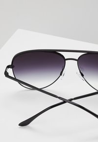 Lunettes de soleil aviateur noires avec des verres dégradés sombres, monture en métal mince, coussinets de nez ajustables et branches courbées croisées sur une surface blanche.
