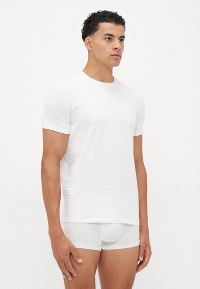 Weißes T-Shirt mit kurzen Ärmeln, das eine strukturierte Oberfläche mit wiederholt aufgedrucktem Markenlogo-Muster aufweist. Kombiniert mit weißer enganliegender Unterwäsche.