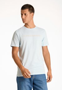 Ljusblå bomulls T-shirt med rund halsringning, med ett beige "COPENHAGEN"-tryck. Bärs med blå jeans. Korta ärmar.