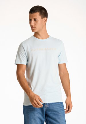 Camiseta de algodón azul claro con cuello redondo, que presenta un gráfico beige de "COPENHAGUE". Llevada con jeans de denim azul. Mangas cortas.