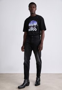 T-shirt noir avec un graphique de palmier coloré et le texte "HUGO", associé à un jean en denim foncé et des bottines noires à talons avec des accents de boucles.