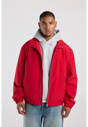 Jonge man die een rood jack met ritssluiting draagt over een grijze hoodie en een blauwe spijkerbroek, staand tegen een effen lichte achtergrond.