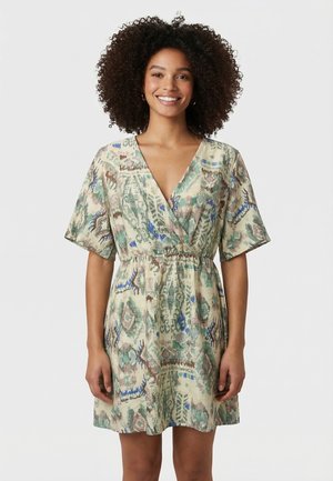 Jeune femme aux cheveux bouclés portant une robe courte à motifs avec un col en V et des manches courtes, debout et souriante devant un fond uni.