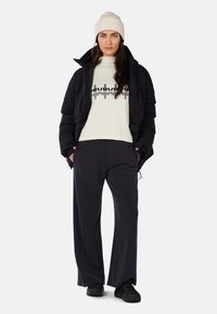 Black puffer jacket, white sweater with black graphic, dark loose-fitting pants, and black shoes. Model wears a light beige knit beanie.