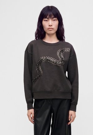 Ung person med langt mørkt hår iført en mørk sweatshirt med et stort slangedesign på forsiden og sorte bukser.