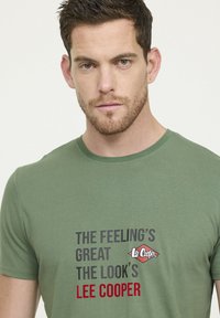 T-shirt en coton vert avec des manches courtes, un col rond et du texte imprimé en noir, gris et rouge, avec un logo sur le devant.
