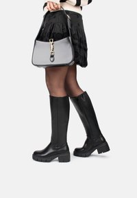 Black knee-high boots with a sleek design, chunky rubber sole, and side zipper. Worn with a black embroidered skirt and a gray handbag.