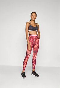 Sutien sport negru cu margini albe, asortat cu colanți roșii cu model floral. Modelul poartă pantofi sport negri, evidențiind un design ajustat.