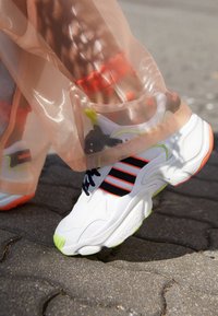 Vit Adidas-sneaker med svarta och orange ränder, neongröna sula accenter, bärs med genomskinlig orange kjol och strumpor med orange hjärt-mönster.