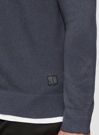 Navy-blauer Pullover mit strukturiertem Strickdesign, gerippten Bündchen und Saum, versehen mit einem angenähten schwarzen Etikett mit weißer Schrift auf der unteren Seite.