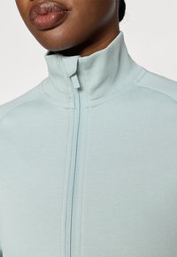 Světlá modrá bunda na zip s vysokým límcem, která má měkkou texturu a jemné šití podél švů.