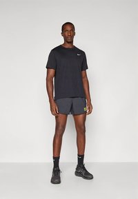 T-shirt atlética preta com mangas curtas e um pequeno logótipo, combinada com calções escuros que apresentam um texto refletor amarelo "BE FAST", sapatos pretos.