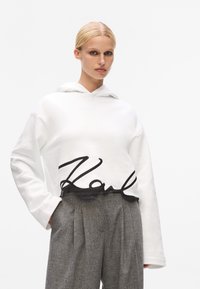 White cropped hoodie with black script logo at the hem, paired with gray pleated trousers. Soft material, relaxed fit, and hooded design.