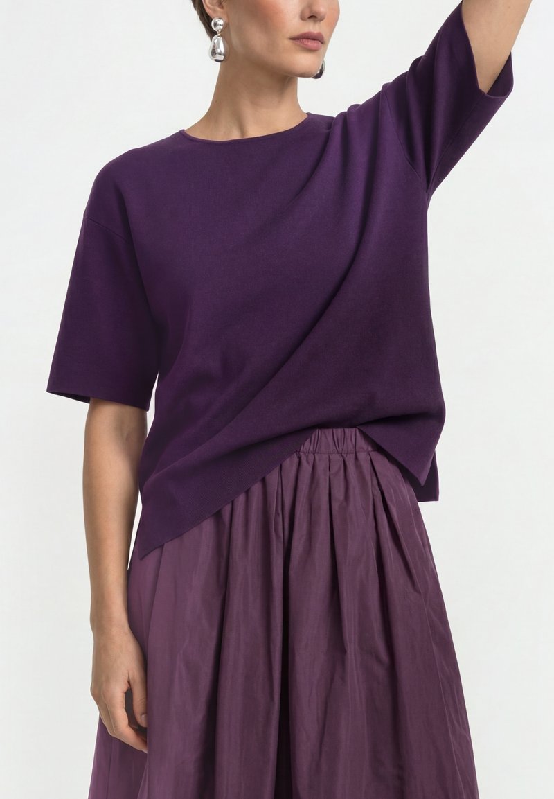 Woman wearing a dark purple short-sleeve top tucked into a pleated purple skirt, accessorized with large silver drop earrings.