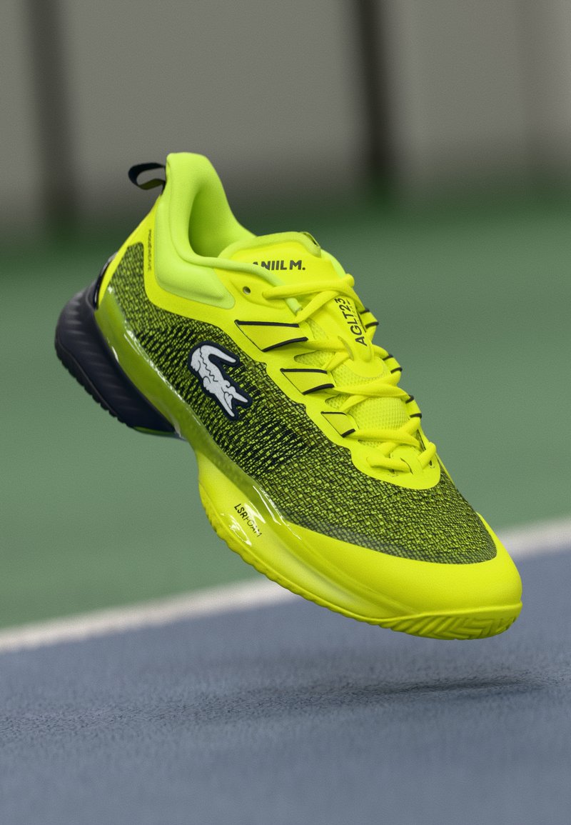 Světlé žluté sportovní boty s texturovaným vrškem z síťoviny, černými akcenty a gumovou podrážkou. Disponují výrazným logem a detaily šněrování.