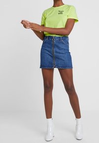 Neonowa zielona koszulka z czarnym napisem, zestawiona z dopasowaną niebieską dżinsową minispódnicą z przednim zamkiem i białymi botkami za kostkę.